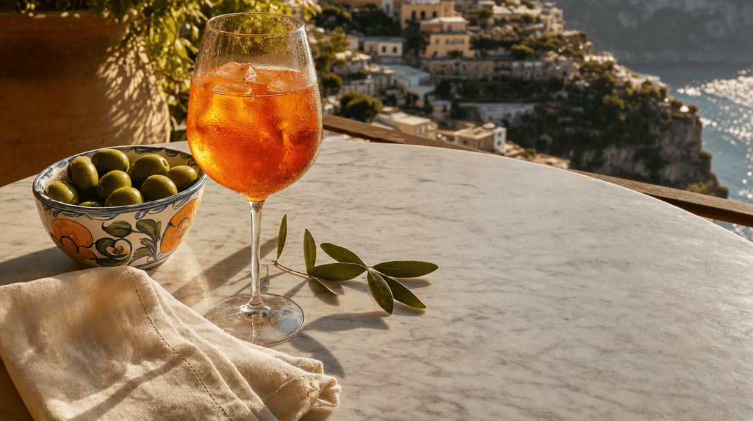 a glass of spritz on a stone parapet overlooking a small cliffside village in afternoon light, olive branches in the foreground