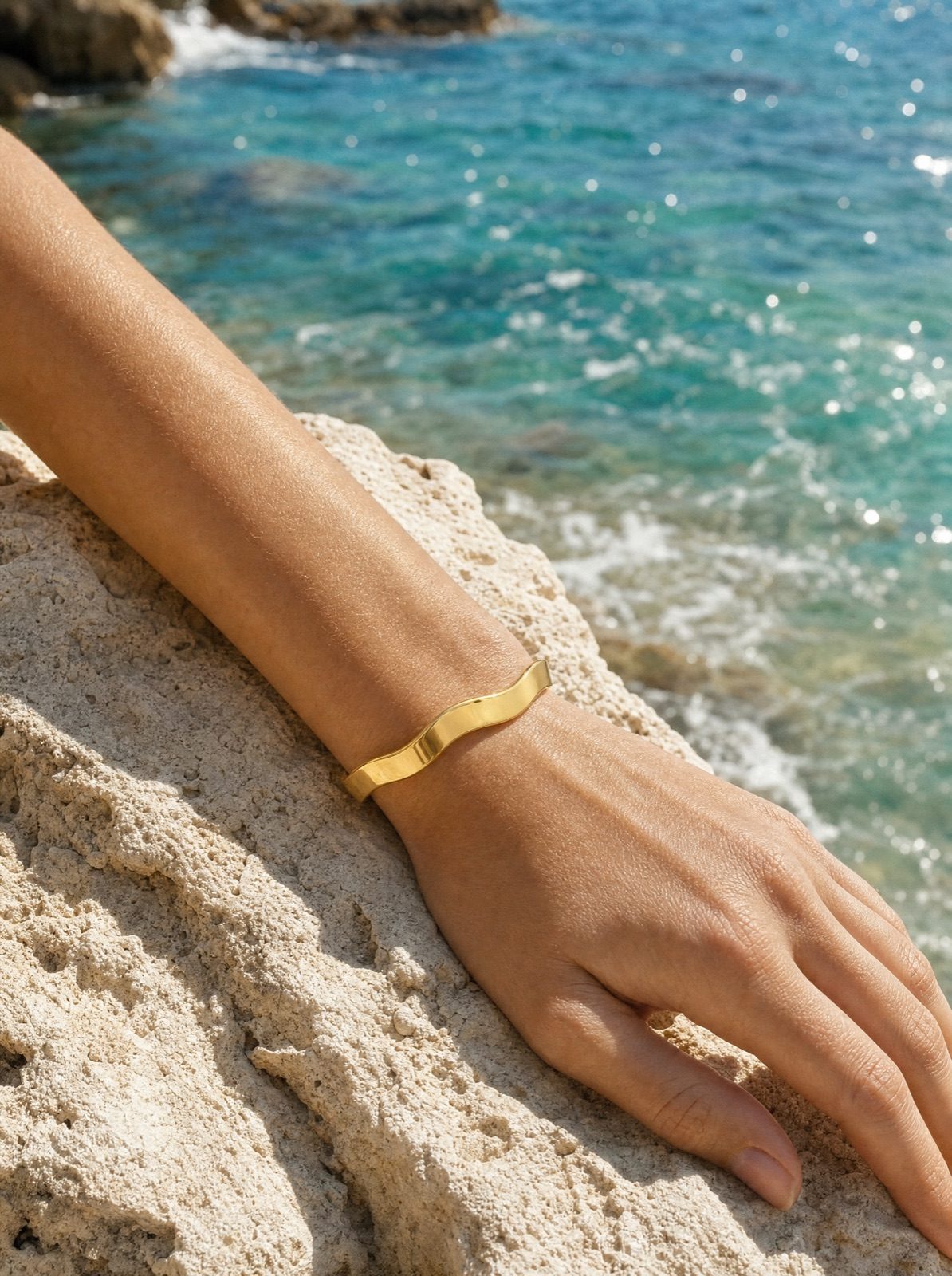 woman wearing the wavy bangle in gold on her wrist, hand resting on a sun-warmed rock above turquoise sea