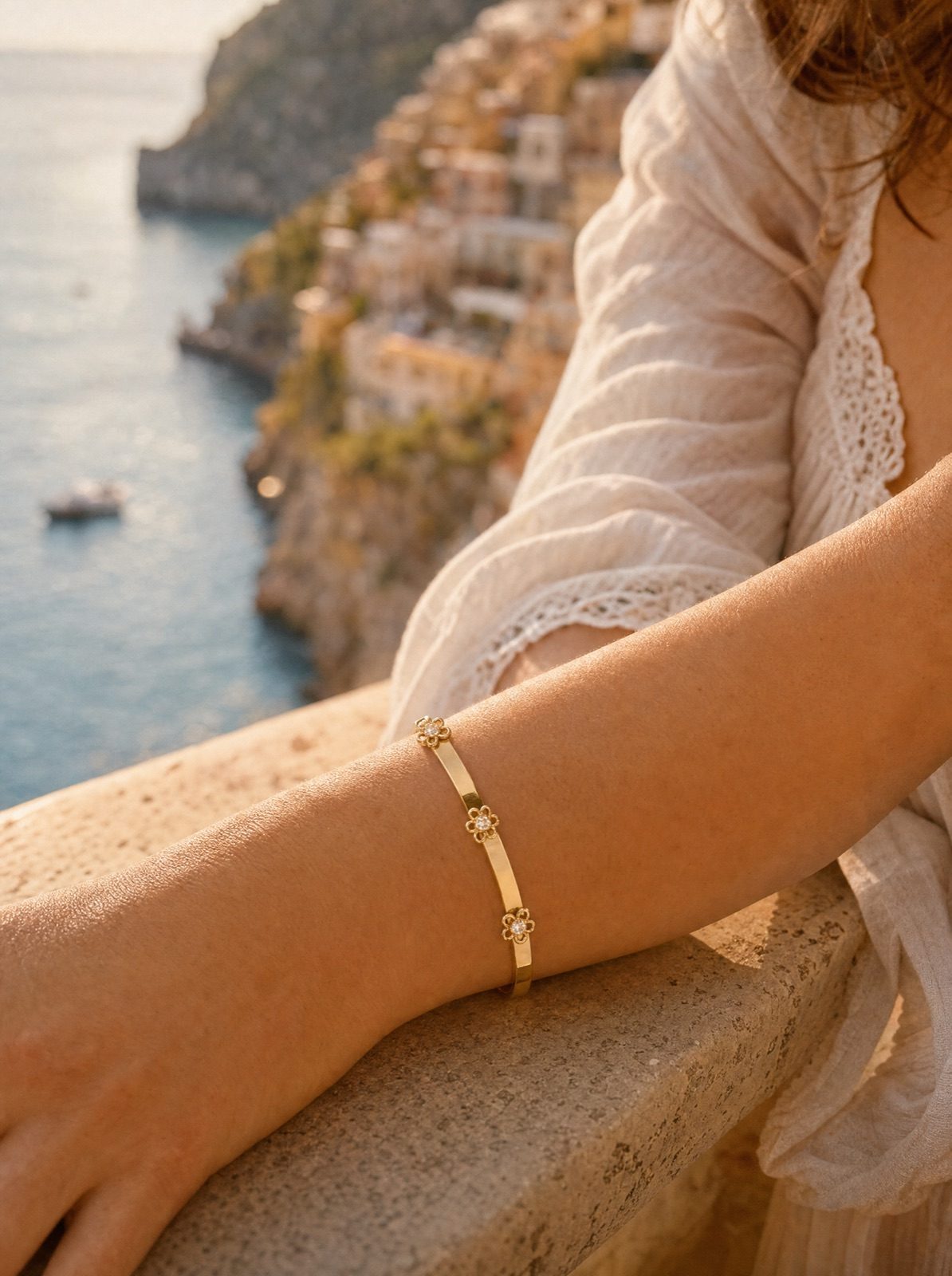 woman wearing the fiore bangle in gold on her wrist, forearm resting on a stone ledge above the amalfi coast