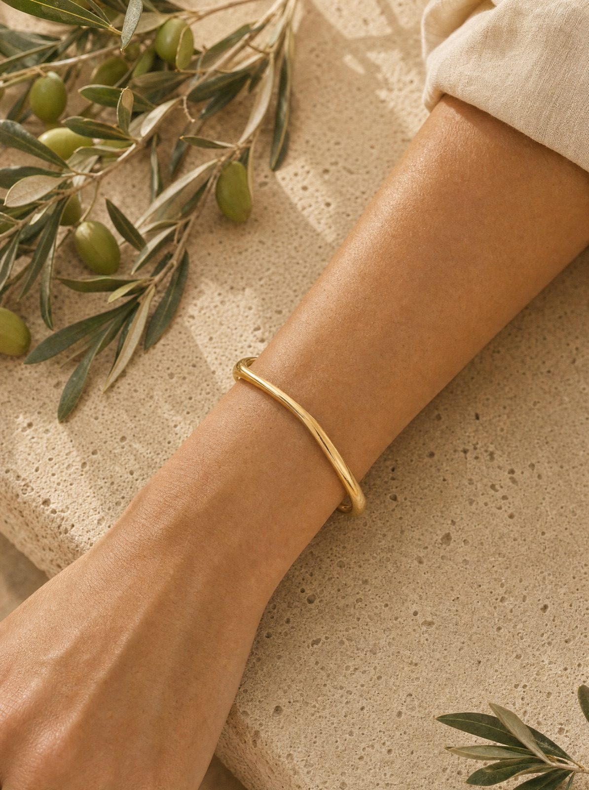 woman wearing the organic cuff in gold on her wrist, forearm on travertine with an olive branch