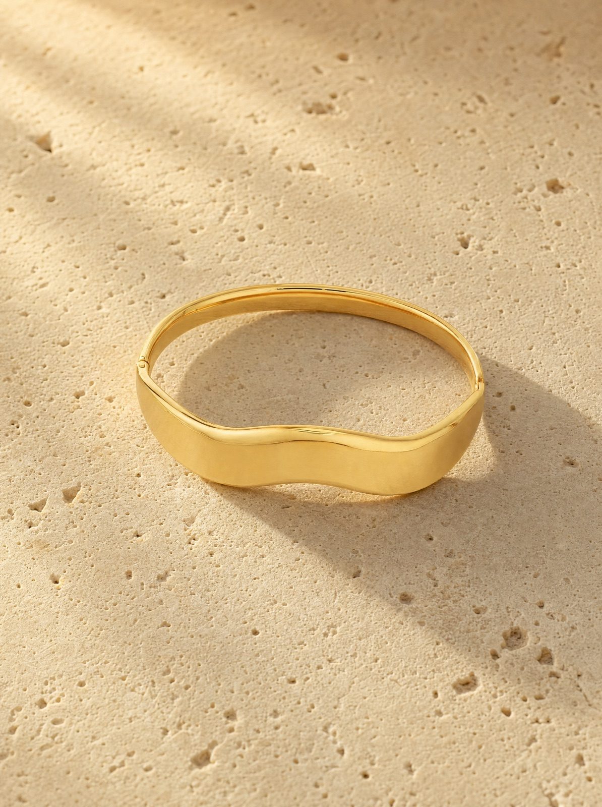 wavy bangle in gold stainless steel on travertine stone in afternoon light