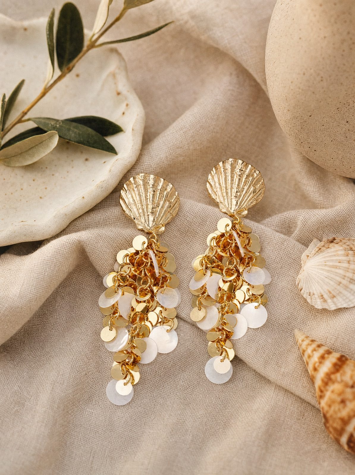 shell earrings with hanging gold sequins on cream linen with an olive branch and seashells