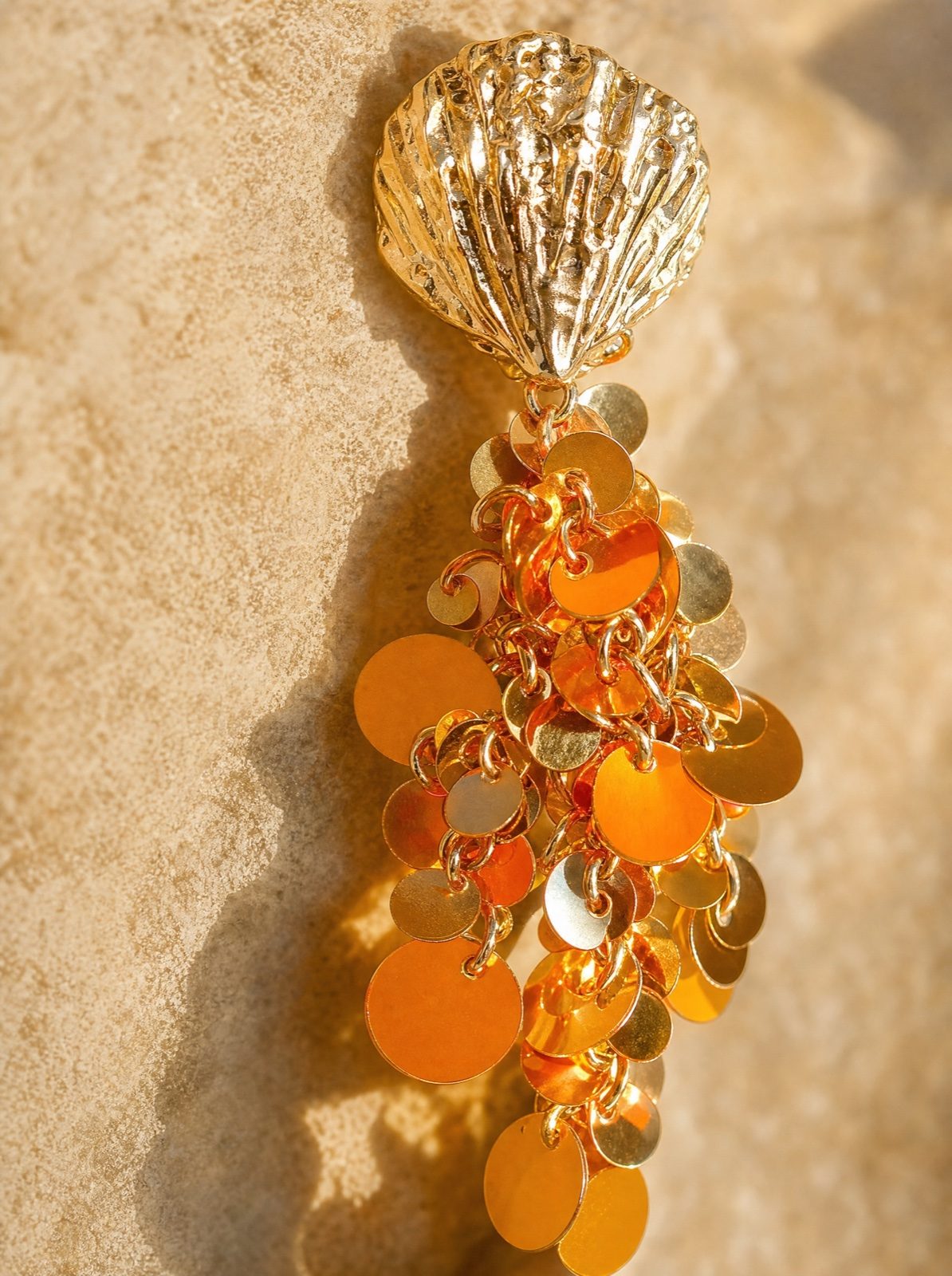 macro detail of a single shell earring with hanging orange sequins on a sandy stone
