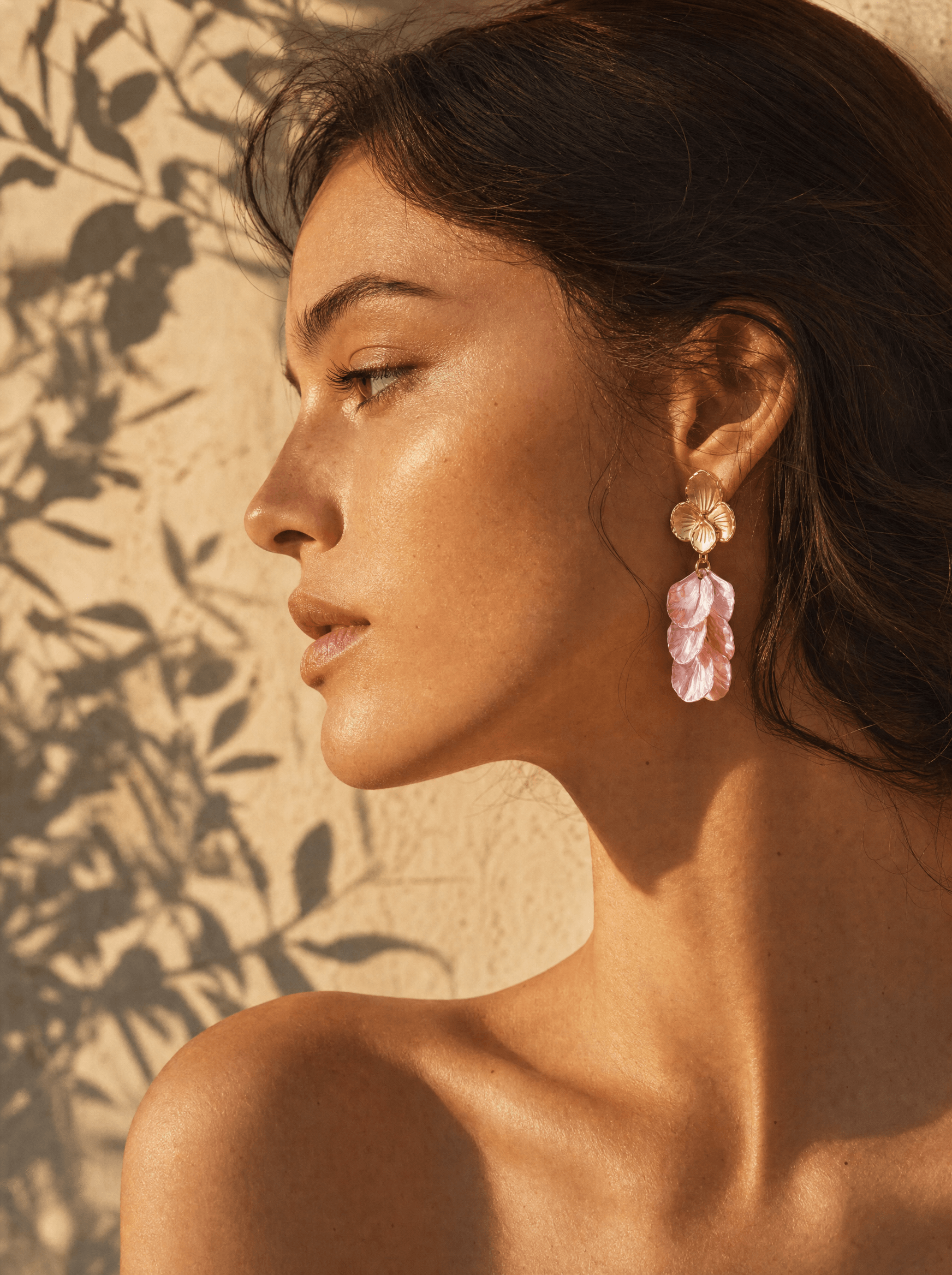 aperi me. — woman in profile wearing the flower earrings in mother of pearl, light pink, with the shadow of olive leaves cast across a warm afternoon wall