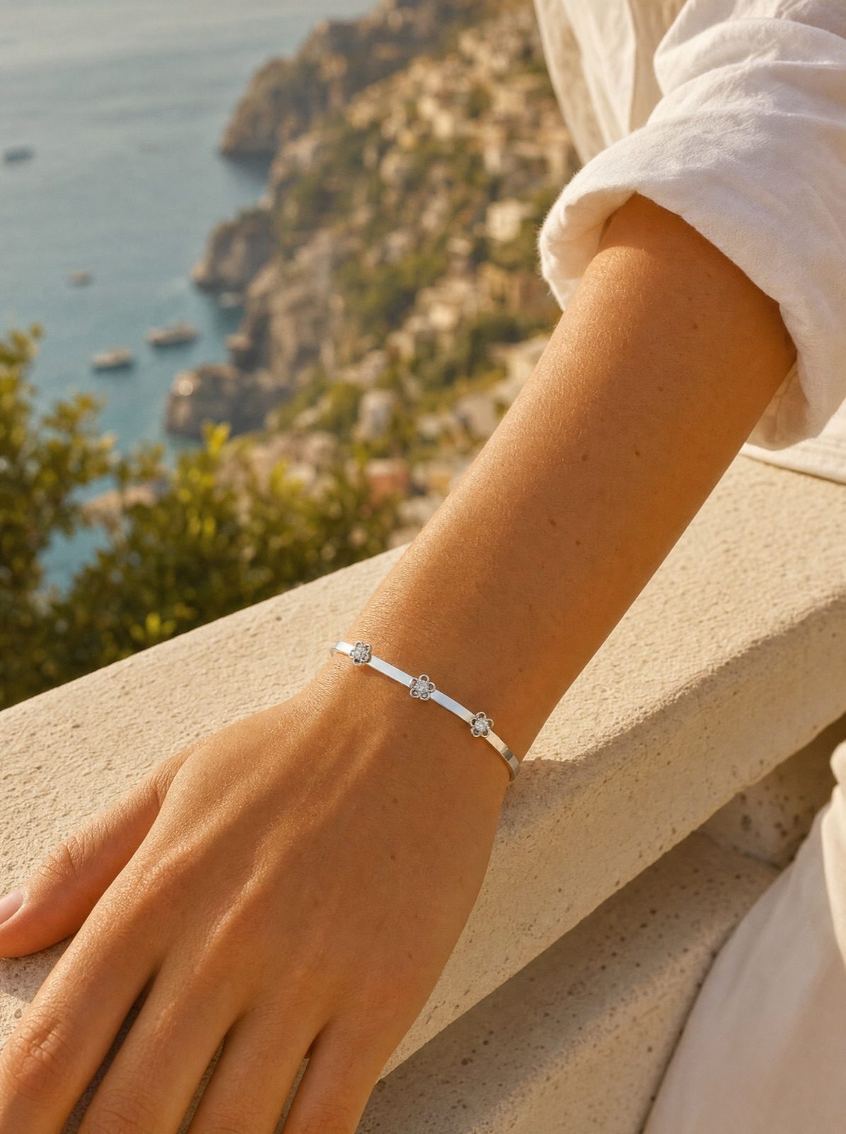 woman wearing the fiore bangle in silver on her wrist, forearm resting on a stone ledge above the amalfi coast